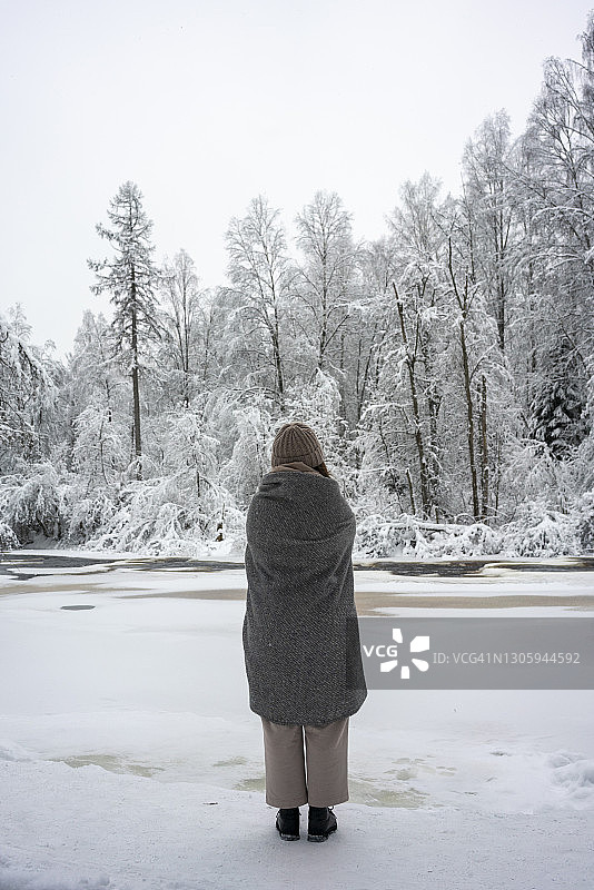 年轻女子裹着毯子站在森林里冰冻的河边图片素材
