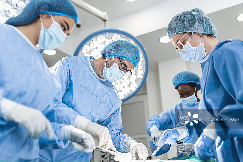在医院手术室对病人进行侵入性手术的专业外科医生，助理和护士的多样化团队。外科医生会说话并使用器械。真正的现代医院。图片素材