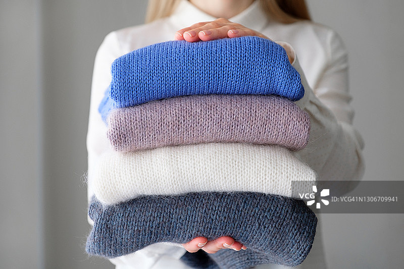 在房间里，一个女人或一个女孩抱着一堆不同颜色的针织品，堆成一堆。冬天和秋天温暖舒适的毛衣慈善。手工制品的储存、护理和洗涤理念。文本空间的副本。图片素材