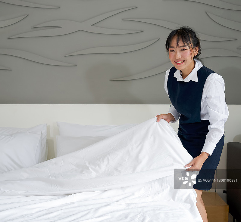 年轻的酒店女服务员微笑着整理床铺。身着蓝色制服的工作人员为酒店客人准备房间。图片素材