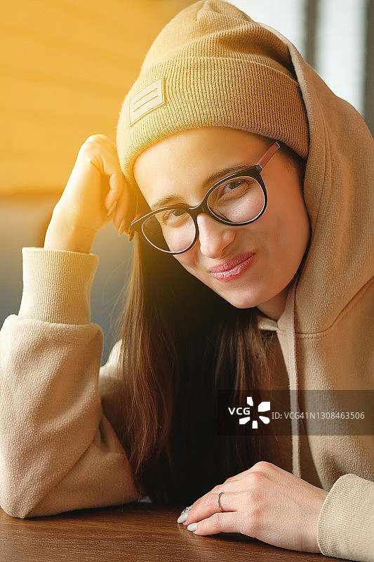 一个美丽的、平静的、体贴的黑发女人，穿着暖和的衣服、帽子和一套西装或针织毛衣在秋冬的街道上。一只手放在她的头旁。一个时尚的年轻女孩或女人的肖像戴眼镜。图片素材