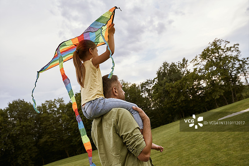 和超级奶爸在一起。一个小女孩坐在她父亲的肩膀上玩彩色的风筝，而站在公园在一个温暖的日子图片素材