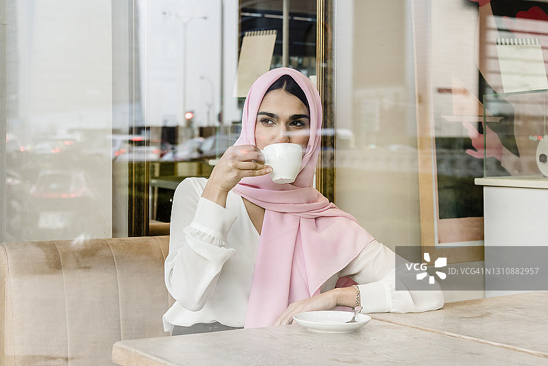 透过窗户看到的肖像是一位年轻的阿拉伯女商人坐在咖啡店里喝咖啡。图片素材