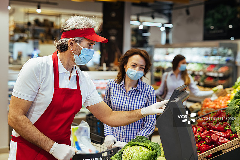 在冠状病毒大流行期间，超市员工帮助客户称蔬菜重量图片素材