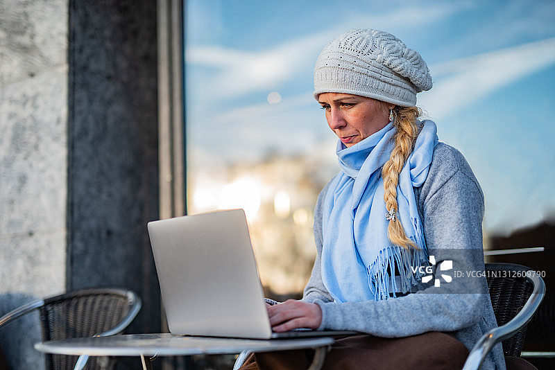 年轻女子穿着暖和的衣服坐在咖啡厅里，在户外使用笔记本电脑图片素材