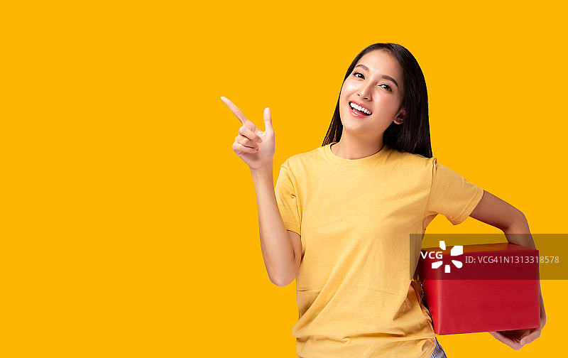 快乐年轻的亚洲女人拿着红色的礼物指向拷贝空间站在黄色的背景美丽的女孩快乐的微笑持有新年礼盒礼物使用为广告黄色的背景图片素材