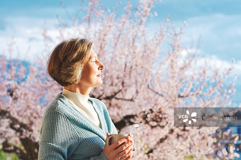 春天的肖像美丽和优雅的中年妇女对粉红色盛开的树，持有一杯咖啡或茶图片素材