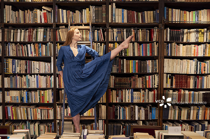 穿着蓝色连衣裙的年轻女子在图书馆的梯子上一只腿保持平衡图片素材