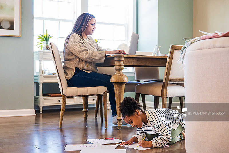 可爱的女孩在纸上画画，而母亲坐在客厅的桌子上使用笔记本电脑图片素材