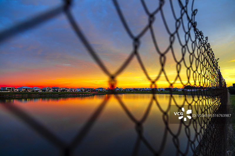 美丽的红色、黄色和橙色的日出透过铁链围栏看向一个湖图片素材