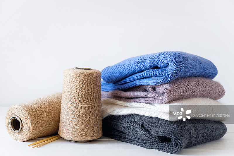 不同颜色的针织品摞在一起，躺在白色的背景上。冬天和秋天暖和的毛衣。在一束束毛线旁边，是编织针。对手工制品的爱好、储存、护理和洗涤的概念。图片素材