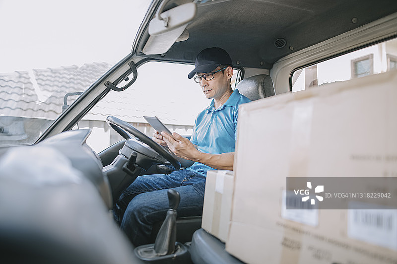亚洲华人成熟男性送货员在送货车的驾驶座上使用平板手机应用查看送货信息图片素材