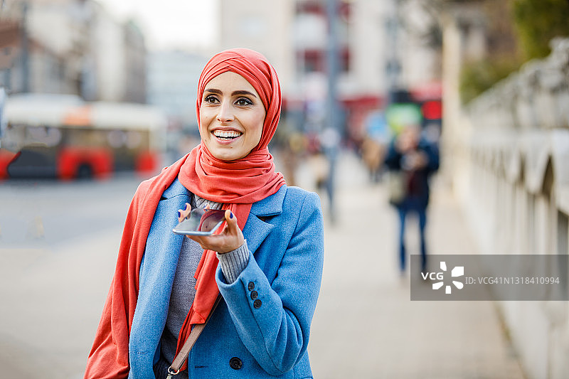 一个穆斯林民族的年轻女子在秋天的一天走在城市的街道上，正在使用手机。图片素材
