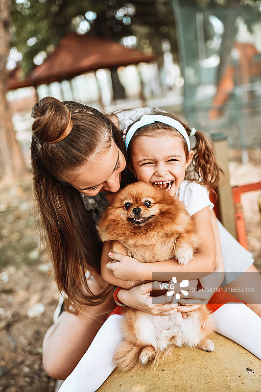 微笑的姐妹拥抱他们可爱的博美犬在操场上图片素材