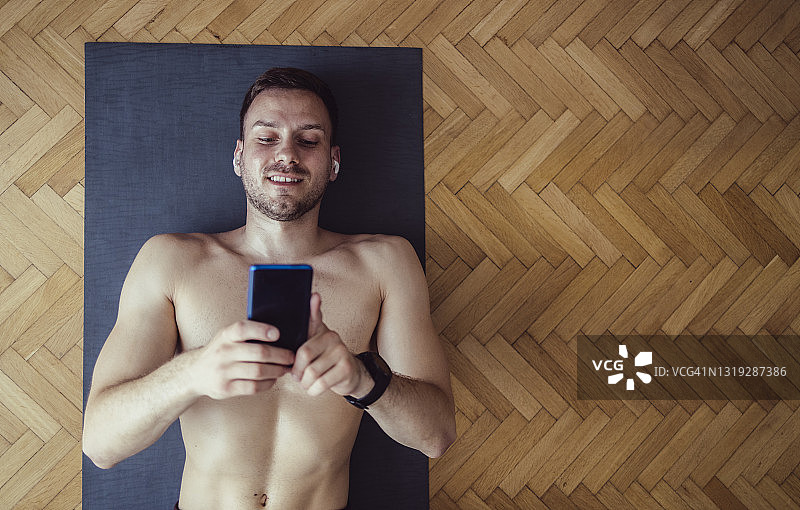带着无线耳机的微笑男子躺在运动垫上，在家锻炼前使用手机图片素材