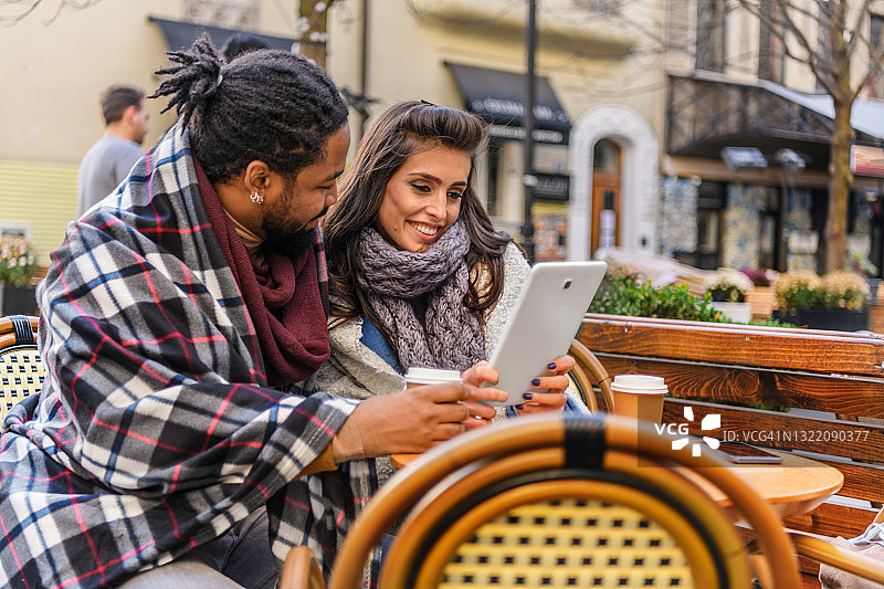 一个年轻的非洲男人和他的妻子正坐在户外咖啡馆的桌子旁，一边喝着咖啡，一边使用着数字平板电脑。图片素材