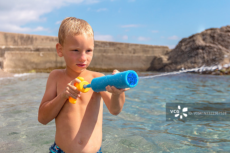 一个晒得黝黑的孩子站在海边的沙滩上，用玩具枪喷出一股水流图片素材