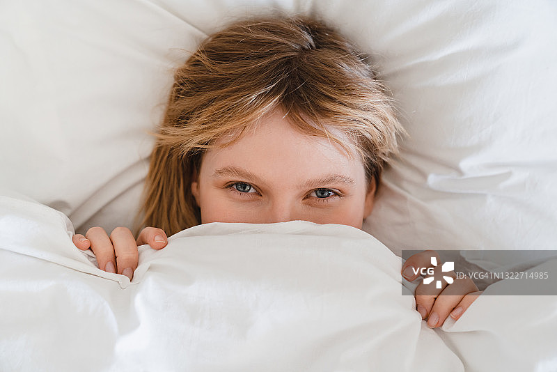 好玩的年轻女子藏在一个舒适的床上的白色毯子亚麻布在早上睡觉和醒来的好心情。平静的床上用品的概念图片素材