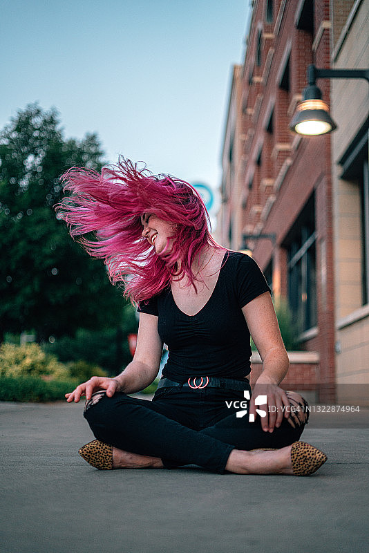 一个美丽的，无忧无虑的活泼时尚的年轻女人与有趣可爱的亮粉色染发坐在人行道上，在夏天在室外的风里甩她的头发图片素材