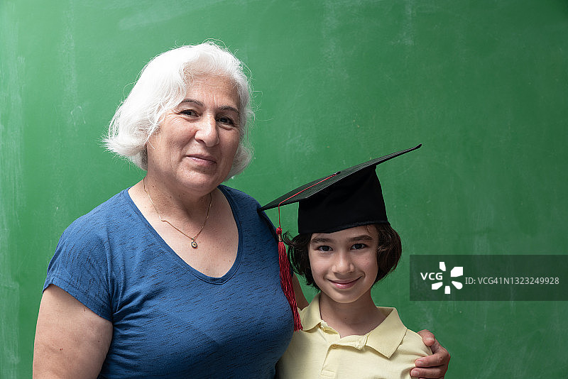 老祖母和孙子在绿色黑板前戴着学位帽的肖像图片素材