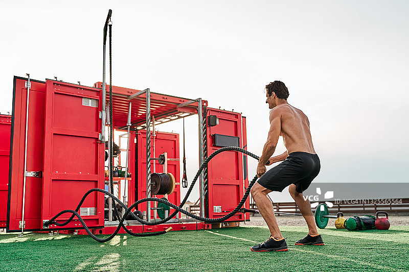 后视图的年轻肌肉男子训练赤膊做交替手臂波浪与战斗绳在一个健身房在海滩上。图片素材