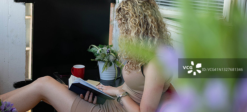 一个卷发的年轻女子坐在家里看书，在阳光明媚的日子里，在她的阳台上享受阅读，享受风景和新鲜的空气，在她的卧室里阅读。图片素材