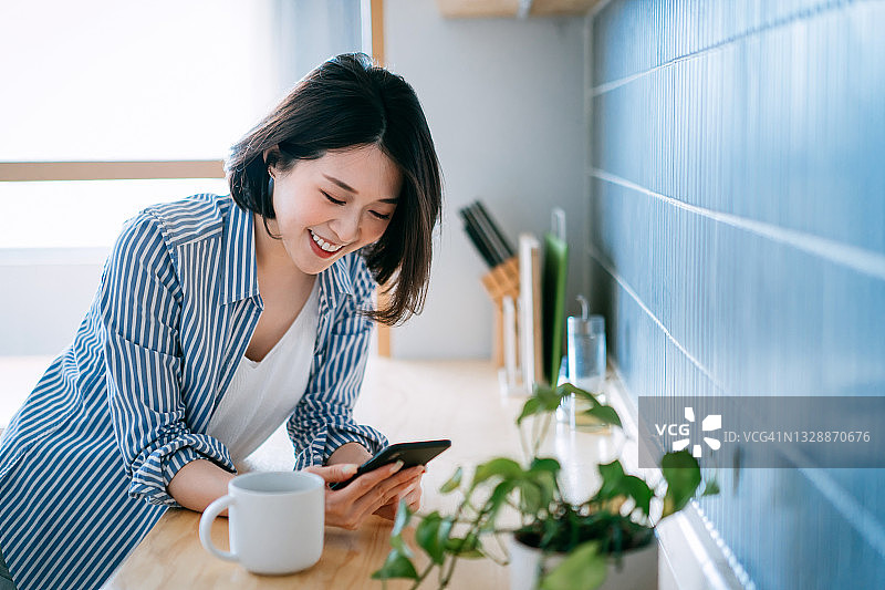 美丽的微笑年轻的亚洲女性使用智能手机，在舒适的家里的厨房里喝一杯咖啡。生活方式与科技理念图片素材