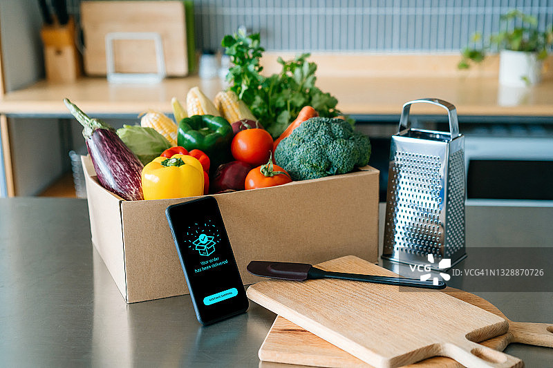 通过智能手机上的移动应用设备在线购买食品杂货，厨房柜台上放着一盒色彩鲜艳、新鲜的有机蔬菜和水果图片素材