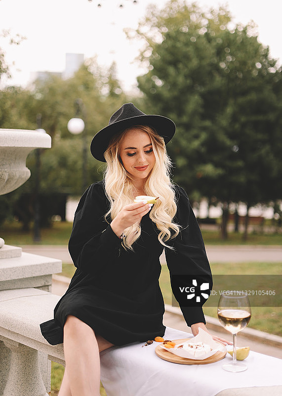 一个美丽浪漫的金发女郎，化着妆，梳着头发，穿着优雅的黑色小礼服，戴着时髦的帽子，坐在公园里的剧院附近的栏杆上，用玻璃杯喝着酒图片素材