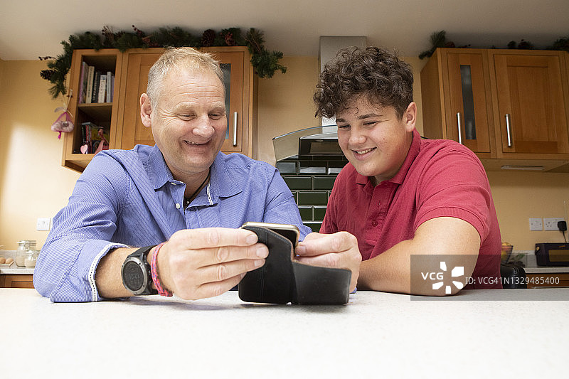 成熟的男人和十几岁的男孩一起看着手机在一个家庭厨房和微笑。图片素材