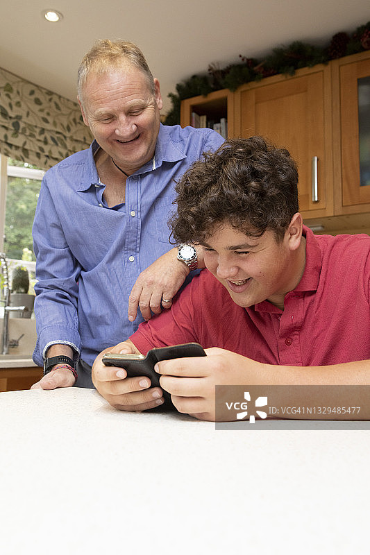 成熟的男人和十几岁的男孩一起看着手机在一个家庭厨房和笑图片素材