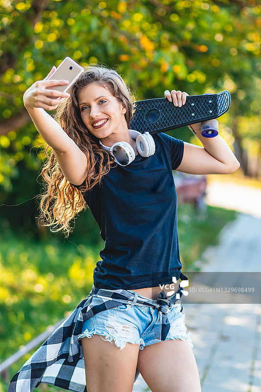 时尚和科技的概念-时尚的年轻女孩在五颜六色的衣服与滑板有乐趣使自拍照在智能手机上图片素材