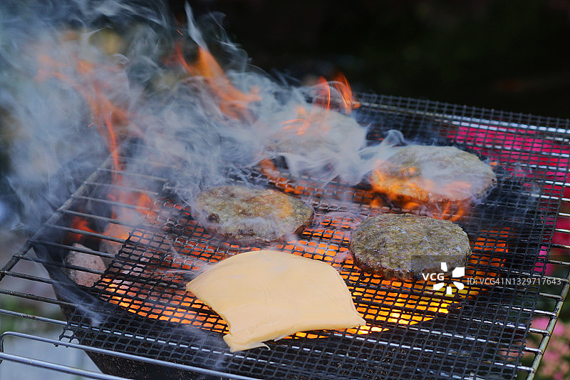 近景拍摄的是便携式的冒烟的火焰，花园里的木炭烧烤，在户外金属烤架上烤的汉堡，在汉堡肉饼上融化的加工过的奶酪片，聚焦前景图片素材