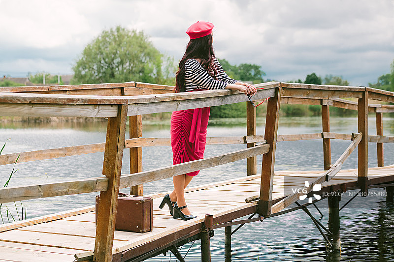 一个年轻的黑发长发女人，戴着红色贝雷帽，戴着黑色眼镜，穿着条纹夹克，红色裙子和黑色鞋子，提着一个复古的手提箱，站在湖上的木桥上，倚着栏杆，望着远方休息图片素材