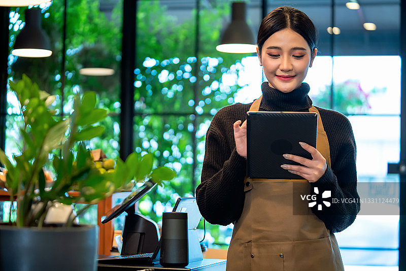 开朗成熟的女服务员拿着她的电子平板电脑，成功的小企业主穿着休闲装，棕色围裙站在咖啡店里。图片素材