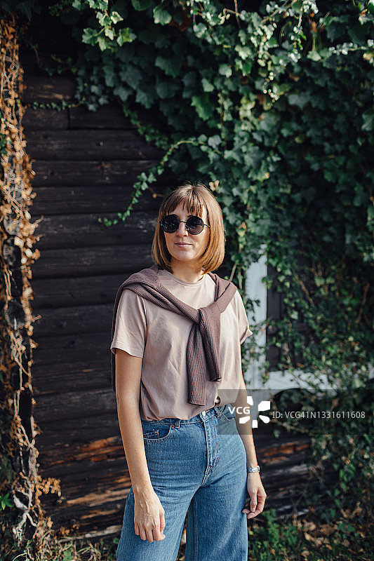 一个年轻的欧洲女孩站在一座木屋旁边，戴着太阳镜看向旁边图片素材