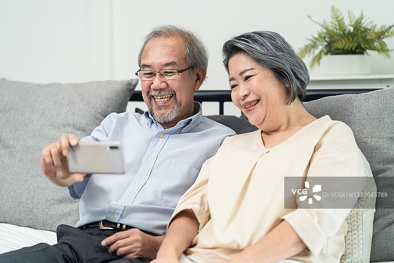 亚洲老年夫妇使用电话远程视频与家人通话。年迈的爷爷奶奶拿着智能手机，在家里的客厅里享受与亲戚和孙子孙女的虚拟在线聊天。图片素材
