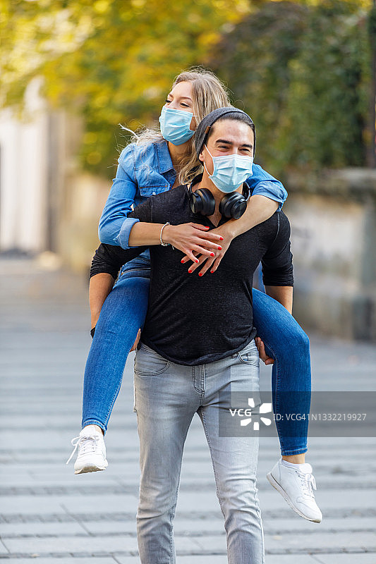 这是一对快乐的恋爱中的年轻夫妇的肖像，他们走在城市的街道上，而在病毒时间里，男人背着他的女朋友。图片素材