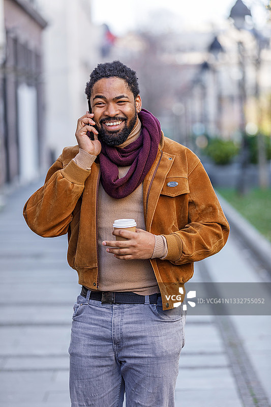 一个英俊的非洲人拿着手机走在城市的街道上，喝着咖啡，用着手机。图片素材