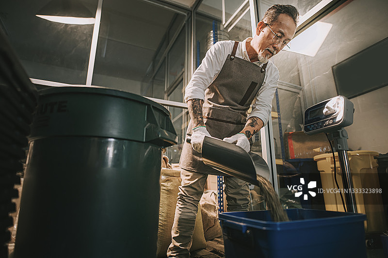 低角度看亚洲华人工作高级男子舀生咖啡豆从桶到称重机和混合它的咖啡烘焙过程在工厂仓库图片素材