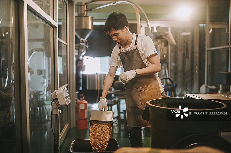 亚洲华人男工人将生咖啡豆从桶中舀到秤机上，并将其混合在工厂仓库的咖啡烘焙过程中图片素材