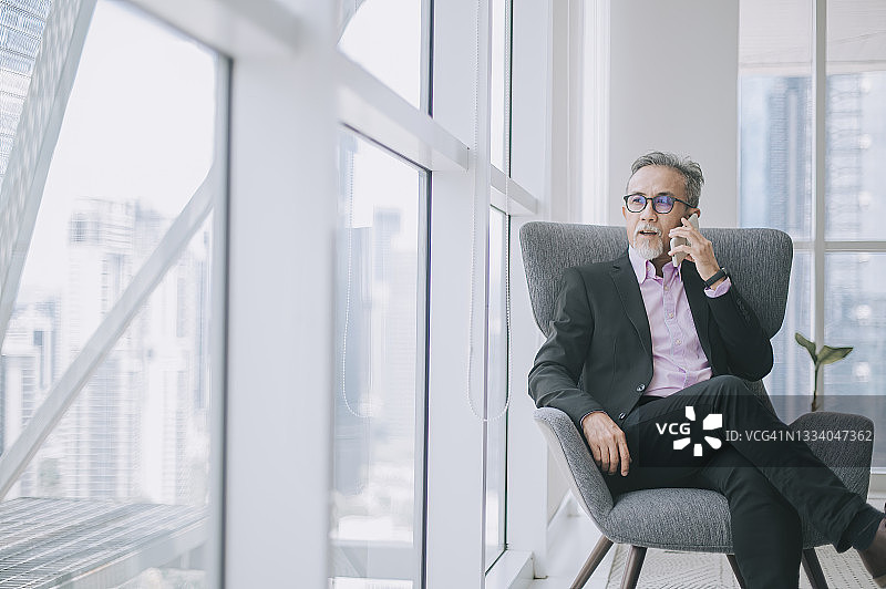 成功的亚洲华裔高级商人穿着西装，盘腿坐在办公室的沙发上听着智能手机图片素材