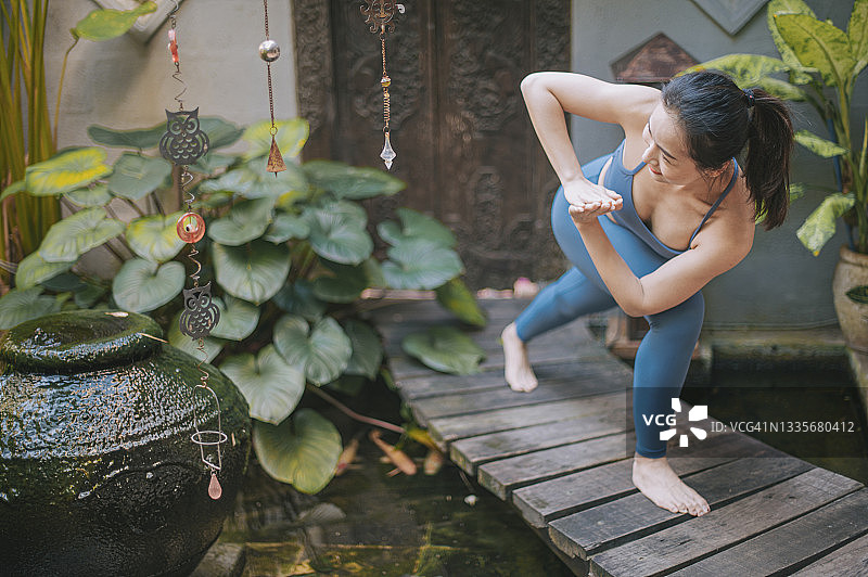 一位亚洲华人中年妇女在前院池塘上方的木桥上练习瑜伽图片素材