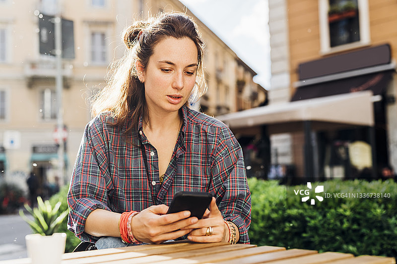 阳光明媚的一天，一名女子坐在路边咖啡馆里使用智能手机图片素材