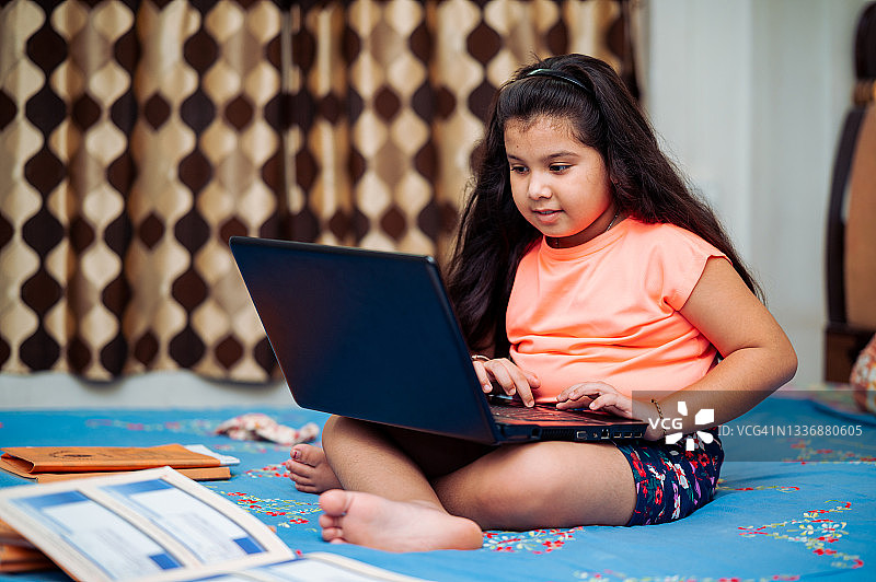 快乐的小女孩在床上用笔记本电脑和智能手机学习。图片素材