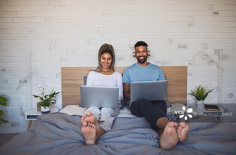 年轻夫妇的照片，笔记本电脑坐在家里的床上，看着相机。图片素材