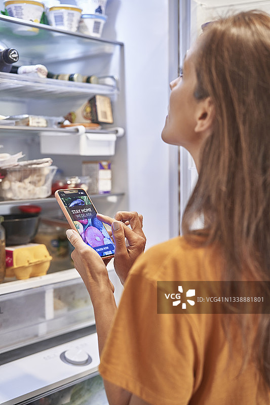 女人一边检查冰箱一边用手机应用订购食品杂货图片素材