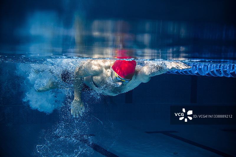 在室内游泳池训练时，身着红色泳帽和护目镜的专业男性游泳运动员的动作和动作的水下视图。健康的生活方式，动力，能量，运动理念图片素材