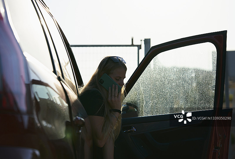 一个金发女孩坐在车门开着的副驾驶座位上用手机打电话的背光。一个金发女人坐在副驾驶上，正在用手机。图片素材