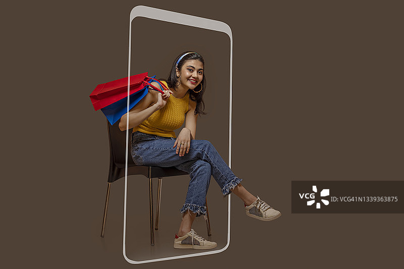 一名年轻女子坐在手机屏幕上，手里拿着五颜六色的购物袋。图片素材
